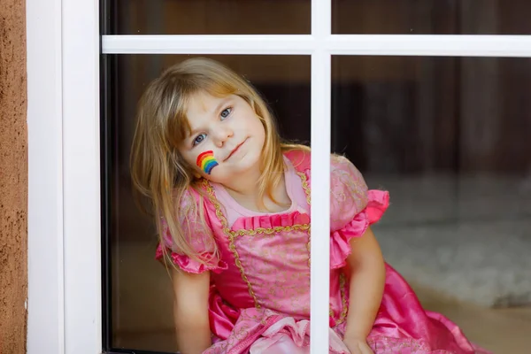 Niedliche einsame kleine Mädchen in Prinzessinnenkleid am Fenster sitzend mit Regenbogen mit bunten Farben im Gesicht während der Coronavirus-Quarantäne. Kinder basteln und bemalen Regenbogen auf der ganzen Welt — Stockfoto