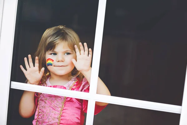 Carino solitario bambina in abito da principessa seduta alla finestra con arcobaleno con colori colorati sul viso durante la quarantena pandemica coronavirus. I bambini fanno e dipingono arcobaleni in tutto il mondo — Foto Stock