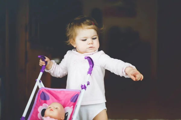 Linda menina adorável fazendo primeiros passos com carruagem de boneca. Criança bonita empurrando carrinho com brinquedo em casa. Filha feliz aprendendo andando e de pé — Fotografia de Stock