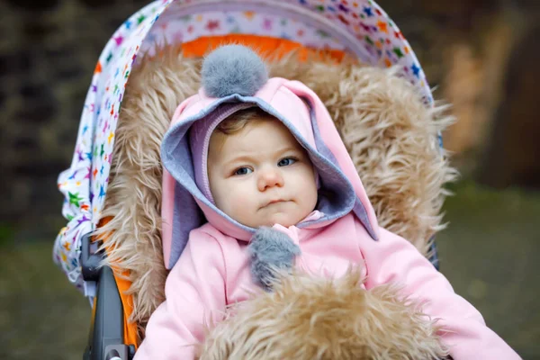 Cute little beautiful baby girl sitting in the pram or stroller on autumn day. Happy smiling child in warm clothes, fashion stylish pink baby coat with bunny ears. Baby going on a walk with parents. — Stock Photo, Image