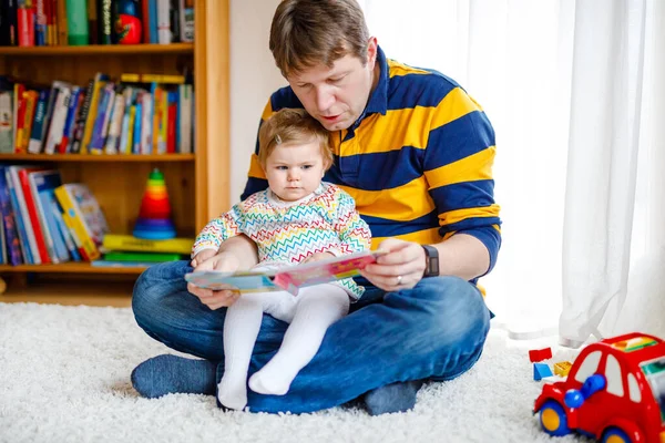 युवा पिता अपनी प्यारी प्यारी बच्ची लड़की के साथ किताब पढ़ रहा है। मुस्कुराते हुए सुंदर बच्चे और आदमी घर में रहने वाले कमरे में एक साथ बैठे। पिताजी को सुनने वाला बच्चा . — स्टॉक फ़ोटो, इमेज
