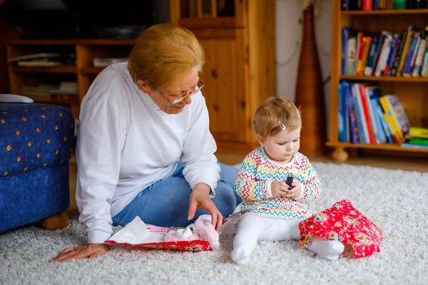 Söt liten småbarnsflicka och mormor leker med leksaker hemma. Förtjusande barn och pensionerad kvinna som packar upp presenter till födelsedagen. Lycklig familj tillsammans — Stockfoto