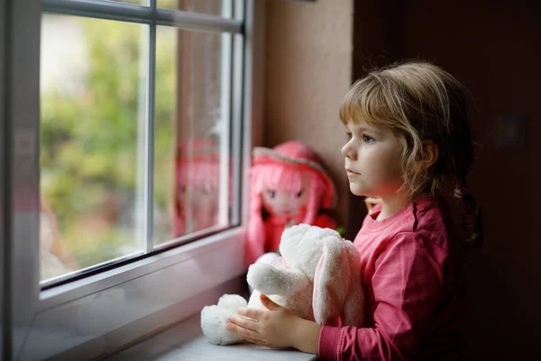 Petite fille mignonne assise à côté de la fenêtre et regardant dehors le jour de pluie. Enfant rêveur avec poupée et jouet doux se sentant heureux. Self isolation concept during corona virus pandemic time. Enfant solitaire. — Photo