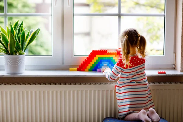 Linda niña pequeña por ventana crear arco iris con bloques de plástico de colores durante la cuarentena pandémica coronavirus. Los niños hicieron y pintaron arcoíris alrededor del mundo como signo. — Foto de Stock