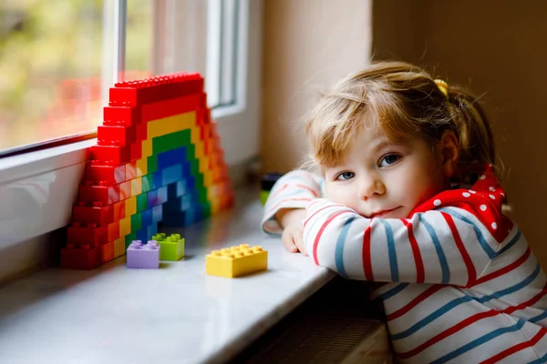 Niedliche kleine Mädchen am Fenster erstellen Regenbogen mit bunten Plastikblöcken während der Coronavirus-Quarantäne. Kinder basteln und malen Regenbogen als Zeichen auf der ganzen Welt. — Stockfoto