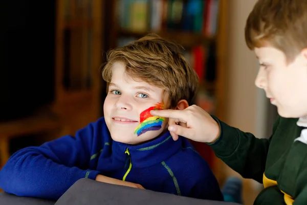 Dos niños con arco iris con colores coloridos en la cara durante la cuarentena del coronavirus pandémico. niño pintar la cara de hermano. Los niños hacen arcoíris en todo el mundo. Hermanos, mejores amigos — Foto de Stock