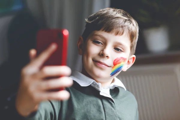 Школьный мальчик с раскрашенной радугой с красочными красками на лице делает селфи со смартфоном. Одинокий ребенок в видеочате или беседе с друзьями во время пандемического карантина коронавируса. — стоковое фото