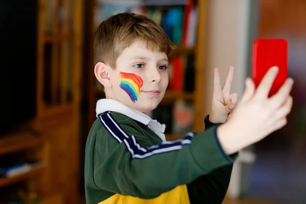 Chłopiec szkolny z malowaną tęczą z kolorowymi kolorami na twarzy zrobić selfie ze smartfonem. Samotne dziecko rozmawiające z przyjaciółmi podczas pandemicznej kwarantanny koronawirusowej. — Zdjęcie stockowe