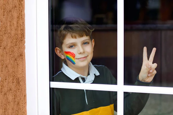 Lonely school kid boy sitting by window with rainbow with colorful colors on face. Child during pandemic coronavirus quarantine. Children make and paint rainbows around the world — Stock Photo, Image