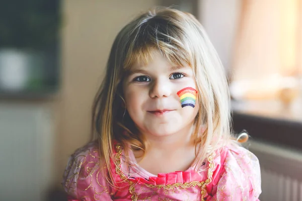 Retrato de linda niña pequeña con arco iris pintado con colores coloridos en la cara. Niño solitario durante la cuarentena del coronavirus pandémico. Los niños hacen y pintan arcoíris en todo el mundo — Foto de Stock