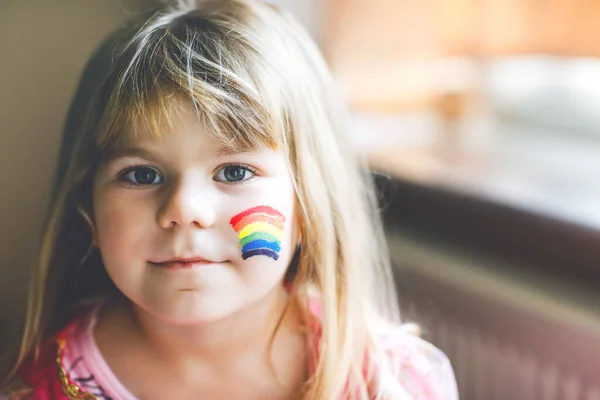 Ritratto di graziosa bambina con arcobaleno dipinto con colori colorati sul viso. Bambino solitario durante la quarantena del coronavirus pandemico. I bambini fanno e dipingono arcobaleni in tutto il mondo — Foto Stock