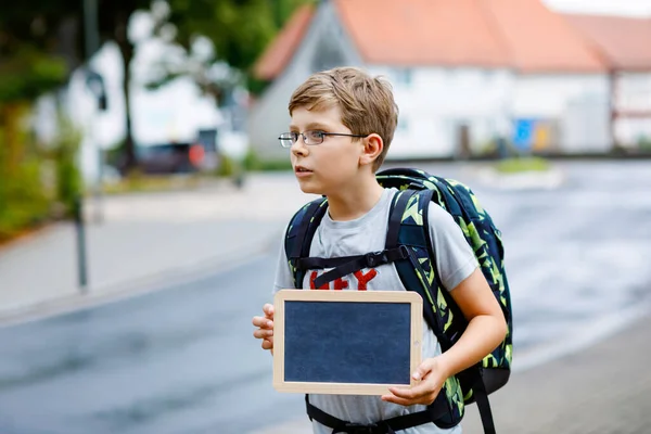 Happy little kid boy with glasses and backpack or satchel. Schoolkid on the way to middle or high school. Child outdoors on the street. Back to school. Kid holding empty chalk desk