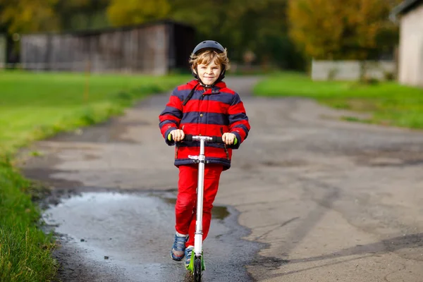 Cute little school kid boy riding on push scooter on the way to or from school. Schoolboy of 7 years driving through rain puddle. funny happy child in colorful fashion clothes and with helmet. — Stock Photo, Image