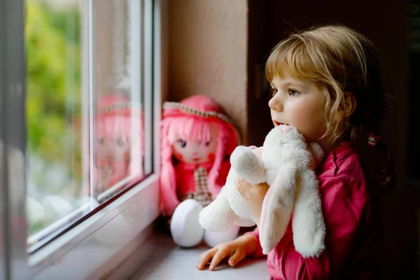 Menina da criança bonito sentado à janela e olhando para fora no dia chuvoso. Criança sonhando com boneca e brinquedo macio sentindo-se feliz. Conceito de auto-isolamento durante o tempo de pandemia do vírus corona. Miúdo solitário. — Fotografia de Stock