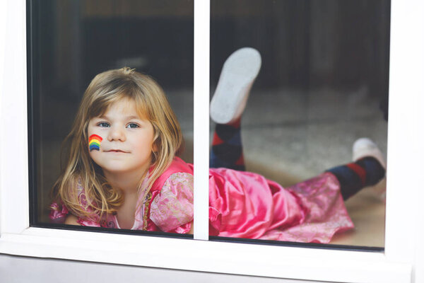 Cute lonely little toddler girl in princess dress sitting by window with rainbow with colorful colors on face during pandemic coronavirus quarantine. Children make and paint rainbows around the world