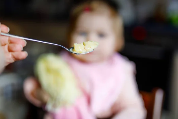 Αξιολάτρευτο κοριτσάκι που τρώει από πουρέ λαχανικών και πουρέ κουταλιού. τροφή, παιδί, σίτιση και άνθρωποι έννοια -χαριτωμένο νήπιο, κόρη με κουτάλι κάθεται σε καρεκλάκι και τρώει στο σπίτι. — Φωτογραφία Αρχείου