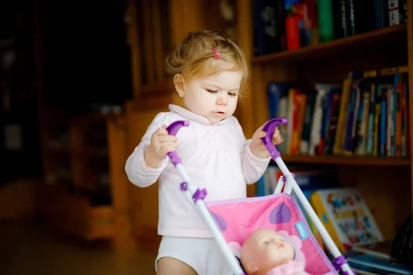 Linda niña adorable haciendo los primeros pasos con carruaje de muñeca. Hermoso niño pequeño empujando cochecito con juguete en casa. Feliz hija aprendiendo a caminar y de pie —  Fotos de Stock