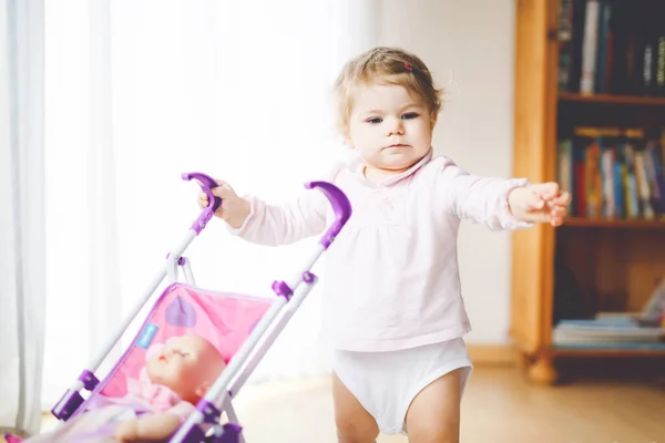 Carino adorabile bambina fare i primi passi con la carrozza bambola. Bellissimo passeggino bimbo che spinge con giocattolo a casa. Felice figlia imparare a camminare e in piedi — Foto Stock