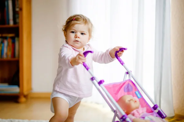 Schattig schattig baby meisje maken eerste stappen met pop rijtuig. Mooi peuter kind duwen kinderwagen met speelgoed thuis. Gelukkige dochter leren lopen en staan — Stockfoto