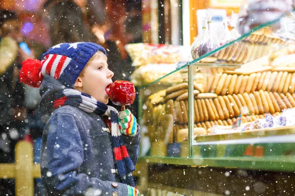 Pequeno menino bonito perto doce stand com pão de gengibre e nozes. Criança feliz comendo em maçã coberta de açúcar vermelho. doce tradicional no mercado de Natal alemão — Fotografia de Stock
