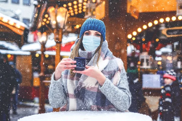 Woman with medical mask drinking hot punch, mulled wine on German Christmas market. People with masks as protection against corona virus. Covid pandemic time in Europe and in the world.