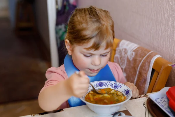 Очаровательная маленькая девочка ест свежий овощной суп на кухне. Счастливый ребенок ест здоровую пищу на обед или ужин. Детское обучение. Дом, детская комната, детская или детский сад — стоковое фото