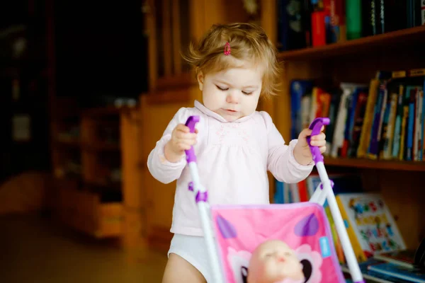 Linda menina adorável fazendo primeiros passos com carruagem de boneca. Criança bonita empurrando carrinho com brinquedo em casa. Filha feliz aprendendo andando e de pé — Fotografia de Stock