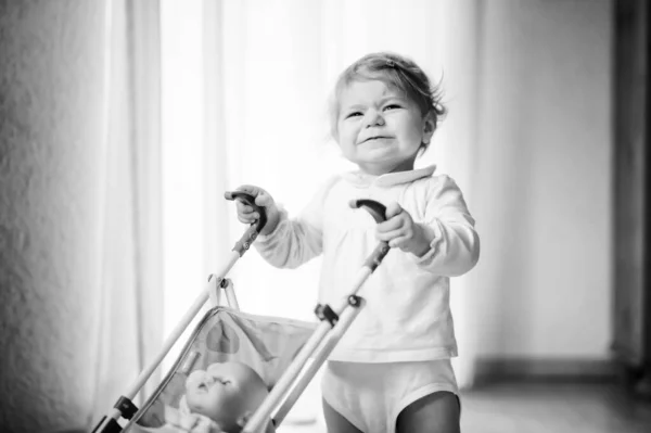 Симпатичная симпатичная девочка делает первые шаги с коляской. Красивый малыш толкает коляску с игрушкой дома. Счастливая дочь учится ходить и стоять — стоковое фото