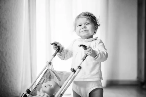 Linda niña adorable haciendo los primeros pasos con carruaje de muñeca. Hermoso niño pequeño empujando cochecito con juguete en casa. Feliz hija aprendiendo a caminar y de pie —  Fotos de Stock