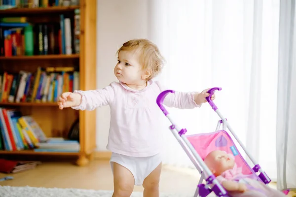 Linda niña adorable haciendo los primeros pasos con carruaje de muñeca. Hermoso niño pequeño empujando cochecito con juguete en casa. Feliz hija aprendiendo a caminar y de pie — Foto de Stock