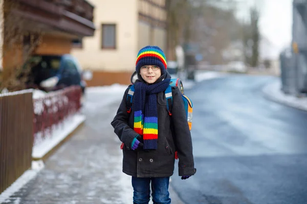 Malý školáček základní třídy, který chodí do školy během sněžení. Šťastné zdravé dítě s brýlemi baví a hrát si se sněhem. S batohem nebo taškou v barevných zimních šatech. — Stock fotografie