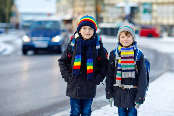 Twee kleine jongens van de basisklas die naar school lopen tijdens de sneeuwval. Gelukkige kinderen die plezier hebben en spelen met de eerste sneeuw. Broers en zussen en vrienden met rugzak in kleurrijke winterkleding. — Stockfoto