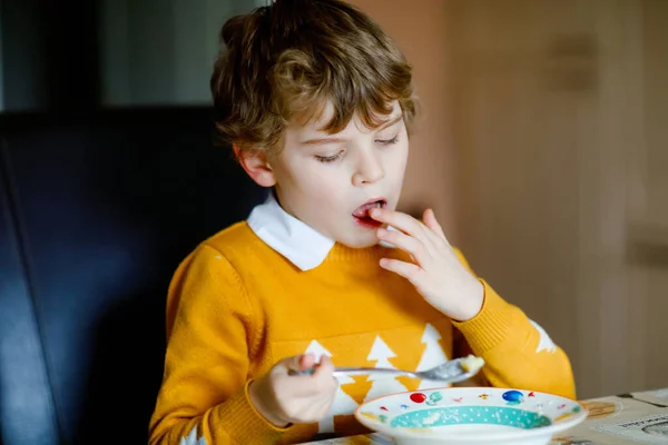 En liten skolpojke som äter grönsakssoppa inomhus. Blond barn i hushållskök eller i skolmatsal. Söt unge och hälsosam mat, ekologisk vegansoppa med nudlar, kyckling och grönsaker. — Stockfoto