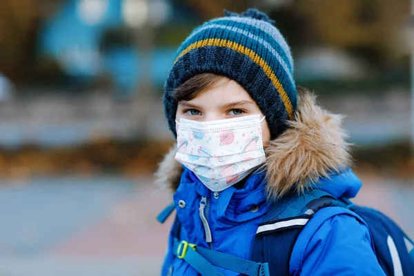 Niño pequeño con máscara médica camino a la escuela. Mochila para niños. Colegial en otoño frío o día de invierno con ropa de abrigo. Tiempo de bloqueo y cuarentena durante la enfermedad pandémica de corona — Foto de Stock