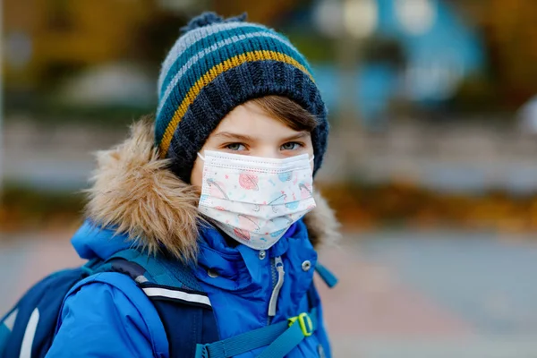 Kleiner Junge mit medizinischer Maske auf dem Schulweg. Schulranzen für Kinder. Schulkind an kalten Herbst- oder Wintertagen mit warmer Kleidung. Sperrung und Quarantäne während der Coronapandemie — Stockfoto