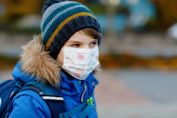 Niño pequeño con máscara médica camino a la escuela. Mochila para niños. Colegial en otoño frío o día de invierno con ropa de abrigo. Tiempo de bloqueo y cuarentena durante la enfermedad pandémica de corona — Foto de Stock