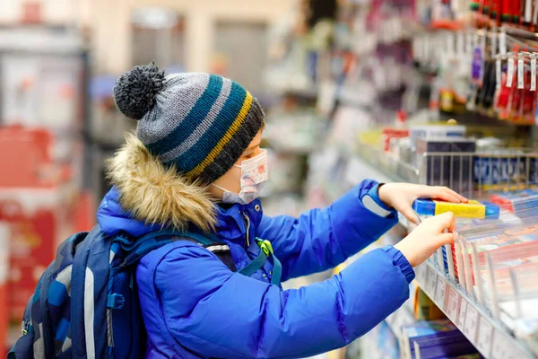 Kleine jongen met een medisch masker koopt schoolspullen in de supermarkt. Kind met rugzak en winterkleding. Schooljongen die potloden koopt. Afsluitings- en quarantainetijd tijdens een corona pandemie — Stockfoto