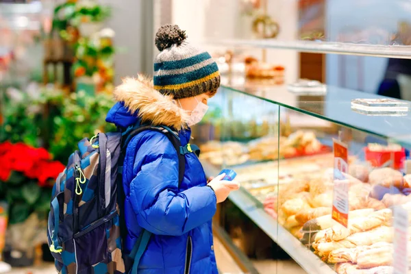 Een jongen met een medisch masker koopt brood en gebak voor de schoollunch in de bakkerij. Kind met rugzak en winterkleding. Schoolkind tijdens vergrendeling en quarantaine tijdens corona pandemische ziekte — Stockfoto