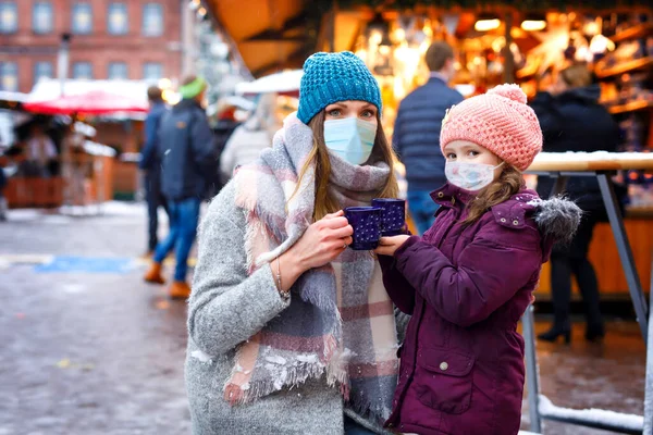 Girl and mother with cup of steaming hot chocolate. Child woman on Christmas market in Germany. People with masks as protection against corona virus. Covid pandemic time in Europe and in the world.