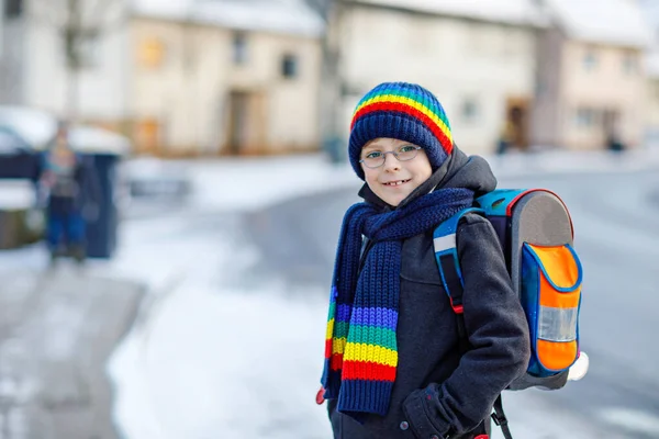 Kleiner Schulbub der Grundschule, der bei Schneefall zur Schule läuft. Glückliches gesundes Kind mit Brille, das Spaß hat und mit Schnee spielt. Mit Rucksack oder Schulranzen in bunter Winterkleidung. — Stockfoto