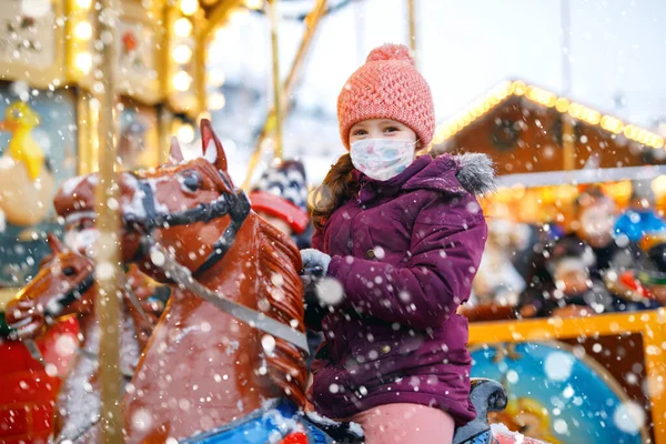 Bambina con maschera medica sul viso cavalcando allegro andare a cavallo giostra rotonda al luna park o al mercato di Natale. maschere come protezione contro il virus della corona. Tempo di pandemia oscura in Europa e nel mondo — Foto Stock