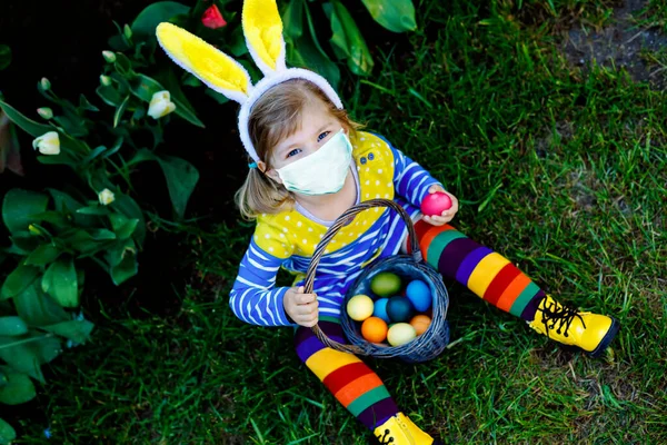 Petite fille tout-petit aux oreilles de lapin qui s'amuse avec la chasse traditionnelle aux œufs de Pâques le jour du printemps, à l'extérieur. Joyeux enfant portant un masque médical. Vacances pendant le temps de confinement de la pandémie de virus corona covid — Photo