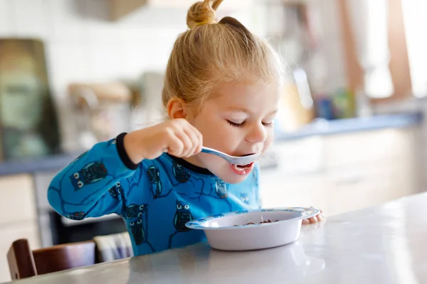 Underbara lilla småbarn flicka äter hälsosamma spannmål med mjölk till frukost. Söt glad baby barn i färgglada kläder sitter i köket och har kul med att förbereda havre, spannmål. Inomhus hemma — Stockfoto