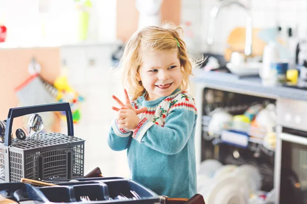 Cute little toddler girl helping in the kitchen with dish washing machine. Happy healthy blonde child sorting knives, forks, spoons, cutlery. Baby having fun with helping housework mother and father. — Stock Photo, Image