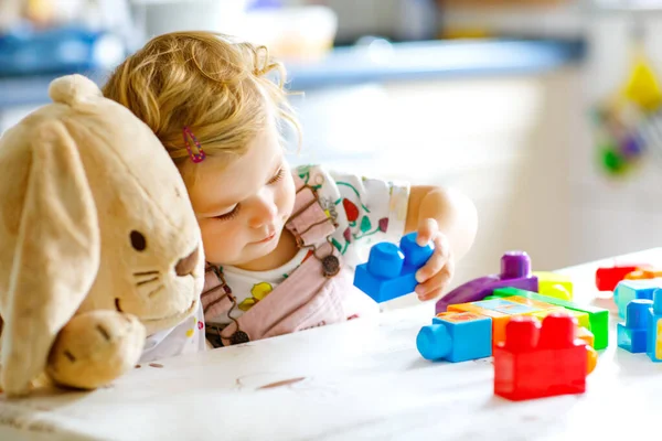 Adorable niña con conejito de peluche favorito jugando con juguetes educativos en la guardería. Feliz niño sano que se divierte con diferentes bloques de plástico de colores en casa. Lindo bebé aprendizaje creación. — Foto de Stock