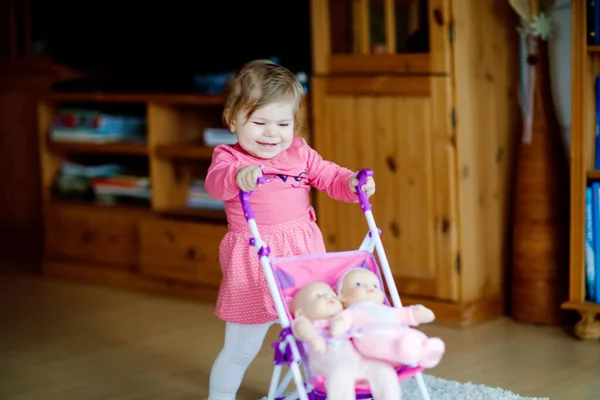 Bayi perempuan manis yang menggemaskan membuat langkah pertama dan mendorong dengan kereta boneka. Anak kecil yang cantik mendorong kereta bayi dengan mainan di rumah. Anak yang bahagia dengan dua boneka — Stok Foto