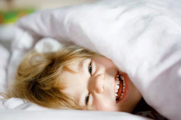 Little toddler girl having fun in bed after wake up. Healthy child jump and play hide and seek with parents. Adorable kid at home, family lifestyle. — Stock Photo, Image