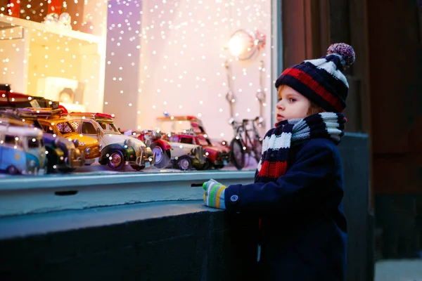 Cute toddler boy looking on car toys in a window on Christmas time season. Fascinated child in winter clothes dreaming and wishing. Window decorated with xmas gifts. Snow falling down, snowfall. — Stock Photo, Image