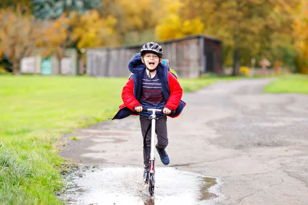 Söt liten skolpojke rider på push scooter på väg till eller från skolan. En skolpojke på sju år som kör genom regnpöl. rolig glad barn i färgglada mode kläder och med hjälm. — Stockfoto
