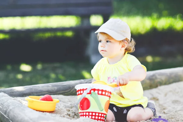 Dışarıda parkta kumlarda oynayan tatlı bir kız çocuğu. Güneşli sıcak yaz gününde güzel bir bebek eğleniyor. Kum oyuncakları ve renkli kıyafetlerle mutlu sağlıklı bir çocuk..